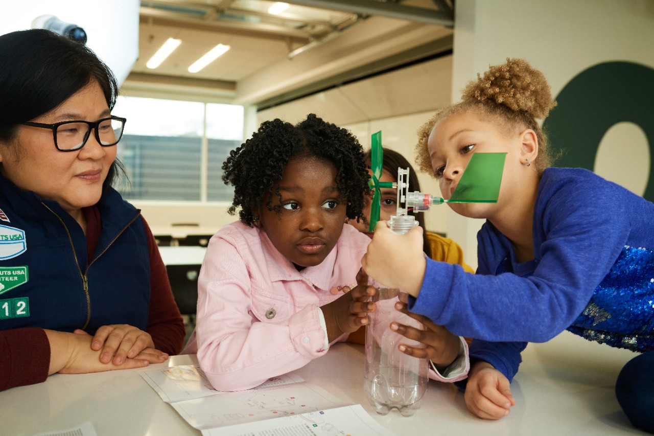 Adult volunteer looking smiling and looking over the shoulder of three Girl Scouts who are coloring at a table. 